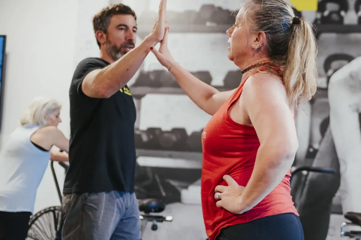Two members share a high-five on the training floor while another member pedals a bike behind them