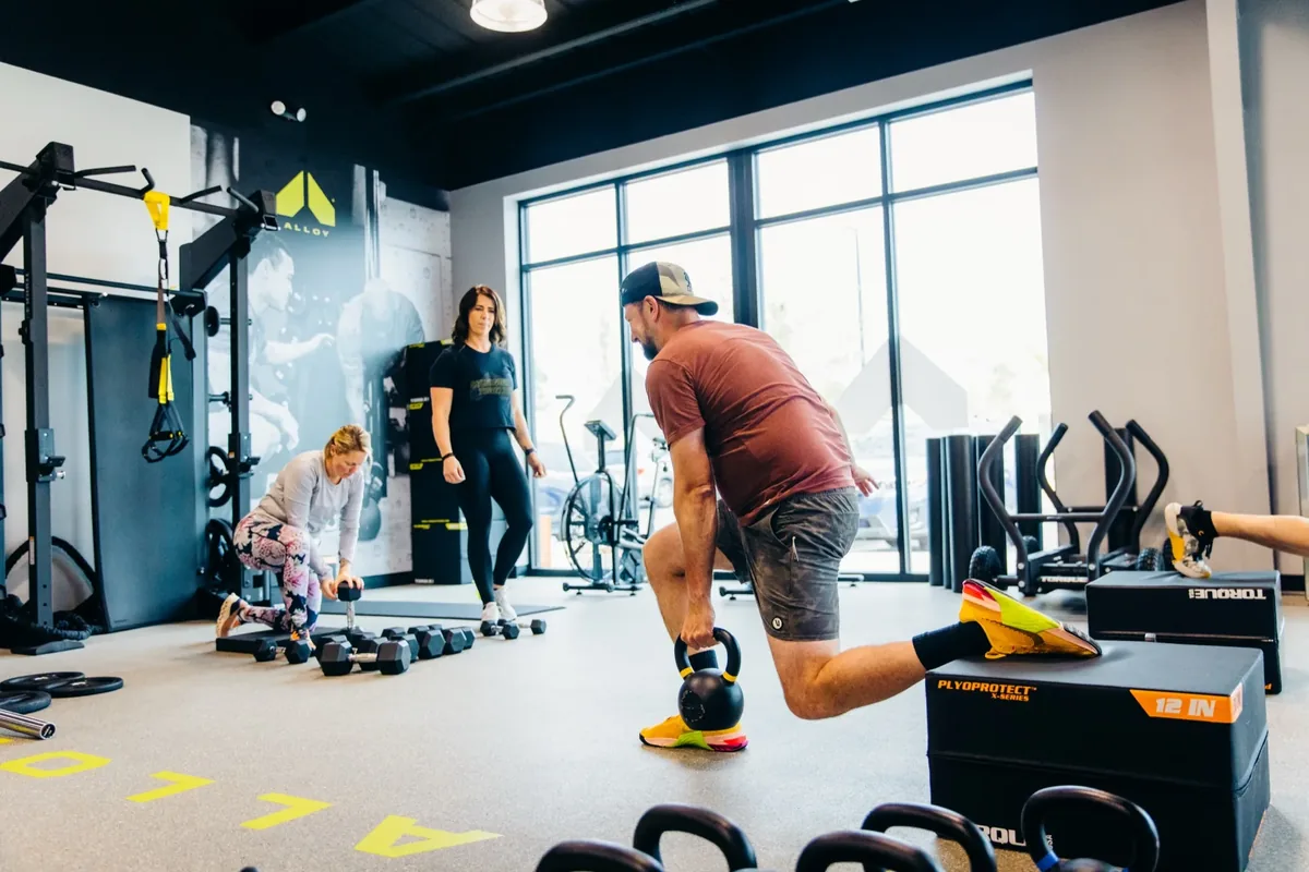 A member drives into a single-leg movement holding a kettlebell while a coach sets up equipment nearby